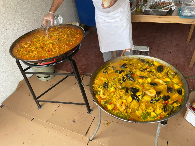 Zwei grosse Paella-Pfannen im Freien, eine behandschuhte Hand gibt etwas hinein